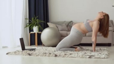 Oturma odasında spor giyim bölümünde hamile bir kadın dizüstü bilgisayarla video dersleri için egzersiz yapıyor. Hamilelik boyunca sağlık ve spor.