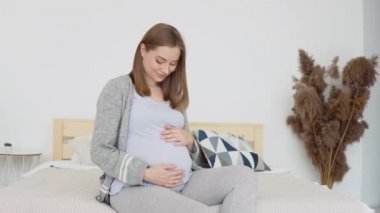 Hamile kadın karnını okşuyor. Çift kişilik yatakta oturan, ev elbiseli hamile bir kadın. İç kısımda kurumuş çiçekler var. Hamileliğin üçüncü üç ayı