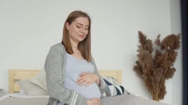 Çift kişilik yatakta oturan, ev elbiseli hamile bir kadın. İç kısımda kurumuş çiçekler var. Hamile kadın karnını okşuyor. Hamileliğin üçüncü üç ayı