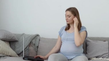 Ev kıyafetleri içinde hamile bir kadın kanepede oturuyor ve dizüstü bilgisayar kullanıyor. Hamileliğin üçüncü üç ayı. Yeni doğan bebeğin doğumu ve bakımı için hazırlık