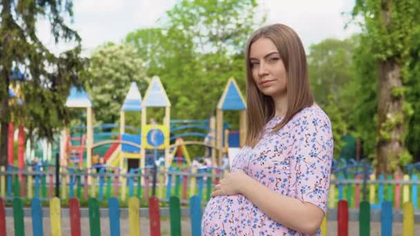 Une femme enceinte dans une robe d'été avec un imprimé floral se dresse sur le fond d'une aire de jeux, où de nombreux enfants jouent activement — Video