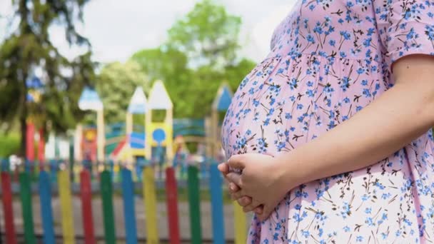 Ciężarna kobieta w letniej sukience z kwiatowym nadrukiem stoi na tle placu zabaw, gdzie aktywnie bawi się wiele dzieci. Widok z bliska brzucha ciężarnej kobiety — Wideo stockowe