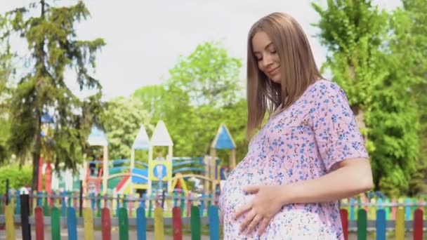 Těhotná žena v letních šatech s květinovým potiskem stojí na pozadí hřiště, kde si mnoho dětí aktivně hraje