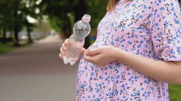 Ciężarna kobieta w letniej sukience z kwiatowym nadrukiem stoi w parku i pije czystą wodę źródlaną wzbogaconą o pierwiastki śladowe. Widok z bliska na brzuch ciężarnych kobiet i butelkę wody — Wideo stockowe