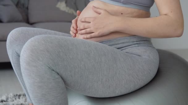 Těhotná žena ve sportovním oblečení sedí na fitballu a drží si ruce na břiše. pohled zblízka na paže a břicho. Zdravotní péče během těhotenství. Fitness pro těhotné ženy — Stock video
