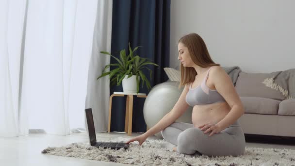 Terhes nő sportruházatban otthon a nappaliban gyakorol videóórákat egy laptoppal. Egészségügyi ellátás és sport a terhesség alatt — Stock videók