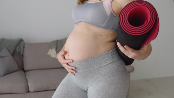 Mulher grávida em sportswear de pé com tapete de fitness nas mãos em casa na sala de estar. Cuidados de saúde e esportes durante a gravidez. Vista de perto da barriga e do tapete — Vídeo de Stock