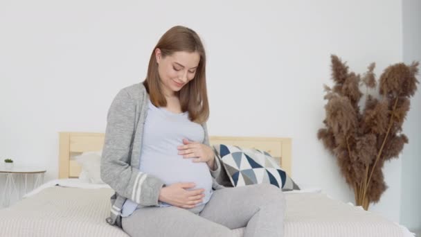 De zwangere vrouw streelt haar buik. Zwangere vrouw in huiskleding zittend op een tweepersoonsbed. Gedroogde bloemen in het interieur. Derde trimester van de zwangerschap — Stockvideo