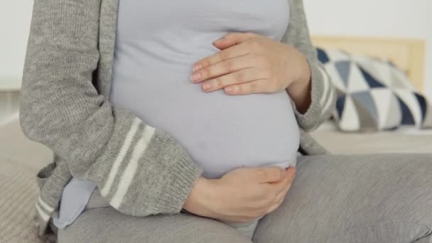 Großaufnahme einer schwangeren Frau in Hauskleidung, die auf einem Doppelbett sitzt. Getrocknete Blumen im Inneren. Die Schwangere streicht sich über den Bauch. Drittes Trimester der Schwangerschaft — Stockvideo