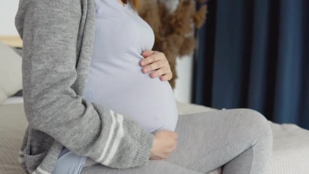 Zbliżenie ciężarnej kobiety w domowych ubraniach siedzącej na podwójnym łóżku. Suszone kwiaty we wnętrzu. Kobieta w ciąży głaska się po brzuchu. Trzeci trymestr ciąży — Wideo stockowe