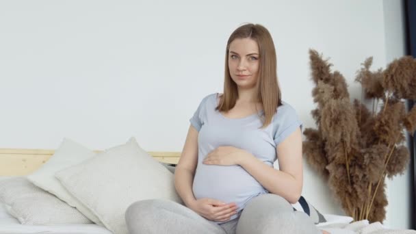 Gravid kvinna i hemkläder sitter på en dubbelsäng. Torkade blommor i inredningen. Den gravida kvinnan smeker magen. Graviditetens tredje trimester — Stockvideo