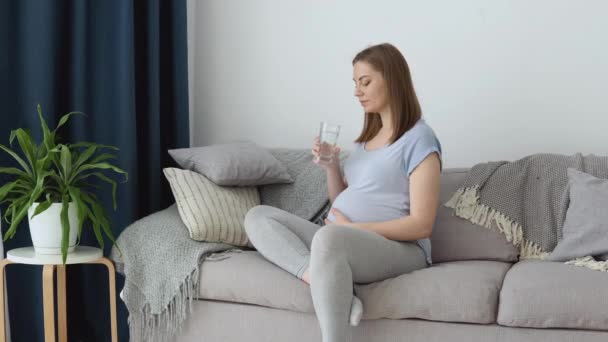 Ciężarna kobieta w domowych ciuchach siedzi na kanapie i pije czystą wodę. Trzeci trymestr ciąży. Zdrowie kobiety w ciąży i jej dziecka — Wideo stockowe