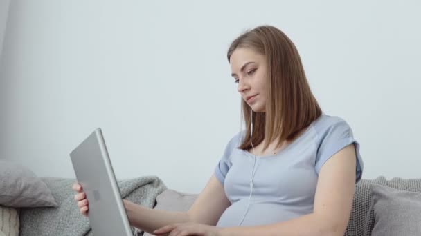 Una mujer embarazada con ropa de casa está sentada en un sofá y usando una computadora portátil. Tercer trimestre de embarazo. Lecciones de fácil parto y cuidado del bebé recién nacido — Vídeo de stock