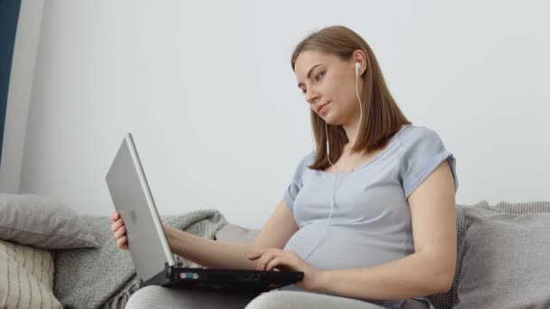 Ciężarna kobieta w domowych ciuchach siedzi na kanapie i używa laptopa. Kobieta w ciąży jako wolny strzelec. Trzeci trymestr ciąży. Lekcje łatwego porodu i opieki nad noworodkiem — Wideo stockowe