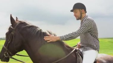 Kasklı, çizgili siyah beyaz gömlekli ve açık renk pantolonlu genç bir adam tarlada siyah bir atın üzerinde oturuyor. Sürücü atın boynunu okşuyor. Profesyonel olmayan bir sürücü dizginleri elinde tutar.
