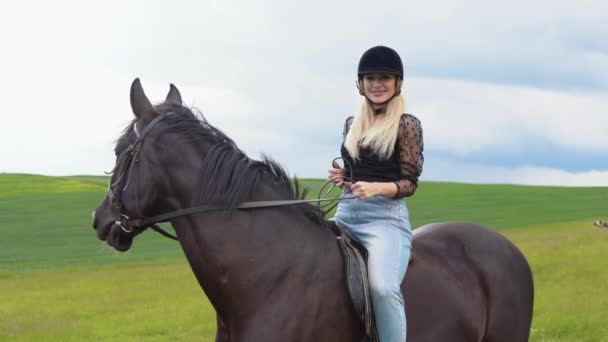 Glamoureuze jonge vrouw in een helm, trendy blouse met doorschijnende weelderige mouwen en lichte jeans zittend op een zwart paard — Stockvideo