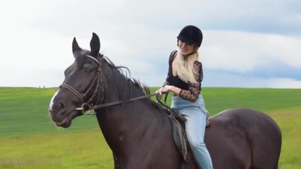 Mujer joven glamurosa en un casco, blusa de moda con mangas exuberantes translúcidas y pantalones vaqueros ligeros se sienta en un caballo negro en el campo. El jinete acaricia el cuello de los caballos — Vídeos de Stock