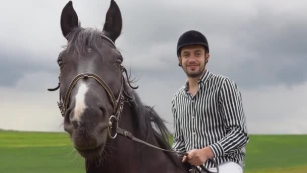 Un joven con casco, camisa blanca y negra a rayas y pantalones ligeros se sienta en un caballo negro en el campo. Terapia ecuestre. Clases de equitación. Un paseo a caballo — Vídeos de Stock