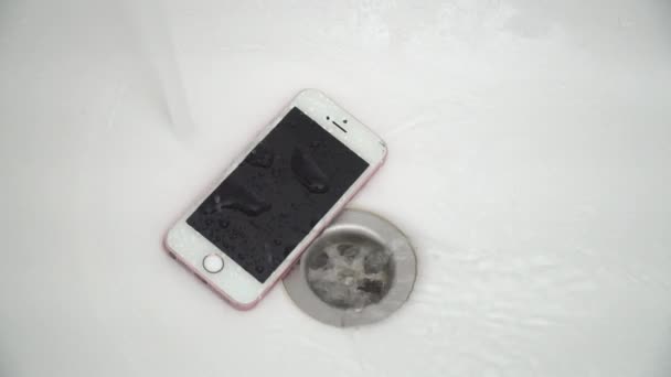 New York, USA - JÚLIUS 1, 2021: Egy rózsaszín-fehér mobiltelefon fekszik egy fehér kád alján, közel a lefolyóhoz. Víz ömlik fentről egy mobiltelefonra. — Stock videók