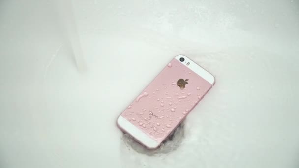 New York, États-Unis - 1er juillet 2021 : Un iPhone rose et blanc est couché sur le fond d'une baignoire blanche près du trou de vidange. L'eau coule d'en haut sur un téléphone portable. — Video