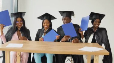 Afro-Amerikan üniversitesinden ya da üniversiteden dört mezun bir masada oturup mavi diplomalarını gösteriyorlar.