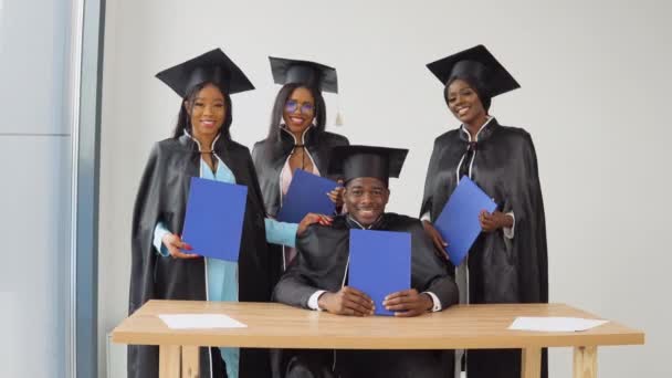 Un hombre está sentado en un escritorio, y tres mujeres están detrás de él. Graduados de una universidad o colegio de nacionalidad afroamericana con diplomas azules en sus manos — Vídeo de stock