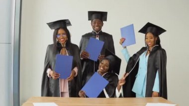 Bir kadın masada oturuyor ve üç sınıf arkadaşı onun arkasında duruyor. Ellerinde mavi diplomalarla Afrika kökenli Amerikan üniversitesinden ya da üniversitesinden mutlu mezun olanlar.