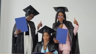 Bir kadın masada oturuyor ve iki sınıf arkadaşı onun arkasında duruyor ve gülümsüyor. Ellerinde mavi diplomalarla Afrika kökenli Amerikan üniversitesinden ya da üniversitesinden mutlu mezun olanlar.