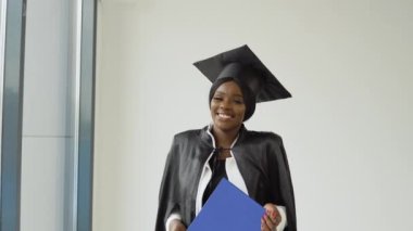 Klasik üstat kıyafeti ve manto giymiş Afrika kökenli bir bayan mezun elinde diplomayla ve gülümseyerek ayakta duruyor. Genç, şık, çekici bir öğrenci.