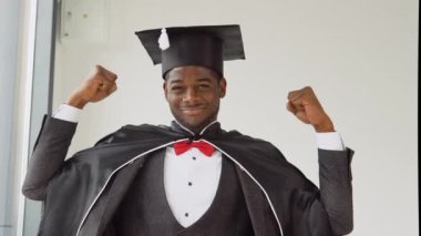 Afro-Amerikan uyruklu genç ve neşeli bir mezun siyah bir cübbe ve sihirbaz şapkası ile kameranın önünde duruyor ve elleriyle gücünü gösteriyor. Samimi gülümseme