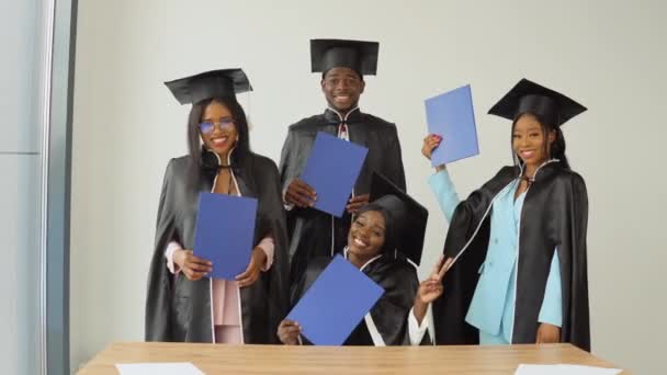 Egy nő ül az asztalnál, és három osztálytárs áll mögötte. Az afro-amerikai állampolgárságú egyetem vagy főiskola boldog végzősei kék diplomával a kezükben — Stock videók