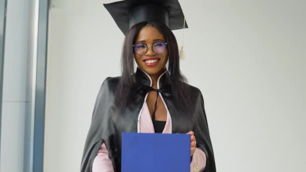 Mujer afroamericana graduada en un traje clásico y el manto de un maestro se levanta con un diploma en la mano y la diversión posando para una foto. Joven elegante estudiante glamoroso — Vídeos de Stock