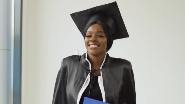 Uma afro-americana graduada em um terno mestrado clássico e manto fica com um diploma em suas mãos e sorri. Jovem elegante estudante glamouroso — Vídeo de Stock