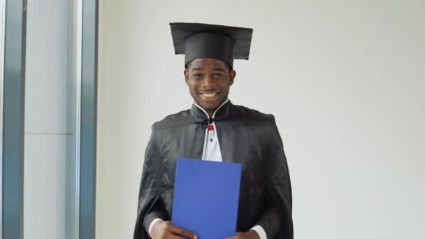 Um jovem e alegre pós-graduado afro-americano está em frente à câmera em um manto preto e um chapéu de mestrado com um diploma de graduação em suas mãos — Vídeo de Stock