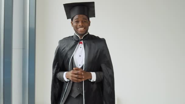 Um jovem graduado alegre de nacionalidade afro-americana fica na frente da câmera em um manto preto e um chapéu de mestres e sorri sinceramente. Sorriso sincero — Vídeo de Stock