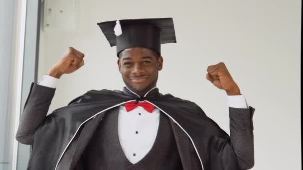 A young cheerful young graduate of African American nationality stands in front of the camera in a black robe and a magisters hat and shows his strength with his hands. Sincere smile — 비디오