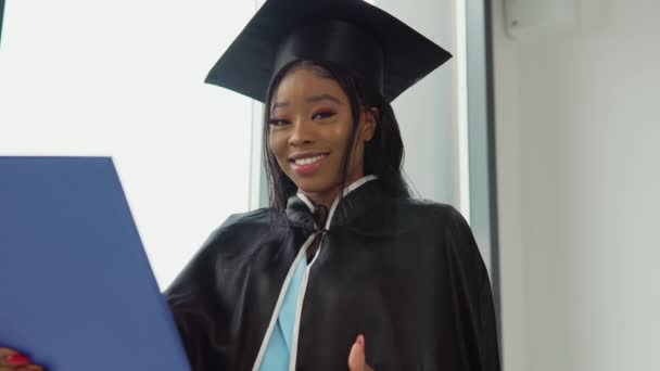 Afroamerykanka kończąca studia w klasycznym garniturze mistrza i płaszcz stoi z dyplomem w rękach i uśmiecha się. Absolwentka Uniwersytetu Medycznego z niebieskim dyplomem w rękach — Wideo stockowe