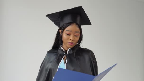 Femme afro-américaine diplômée en costume classique et manteau d'un maître se tient avec un diplôme à la main. Le diplômé regarde le diplôme, puis regarde la caméra et sourit. — Video