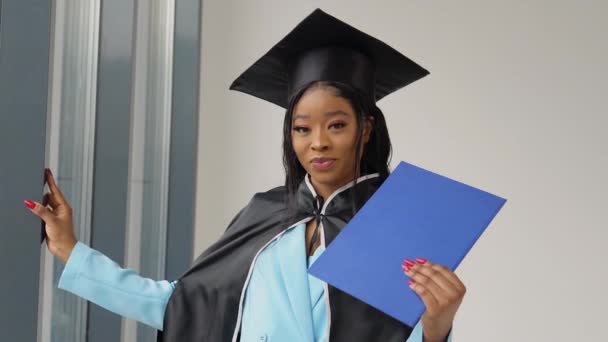 Una mujer afroamericana graduada en un traje y manto de maestros clásicos está junto a la ventana con un diploma en sus manos y sonríe. Una graduada de la universidad con un diploma azul en sus manos — Vídeo de stock