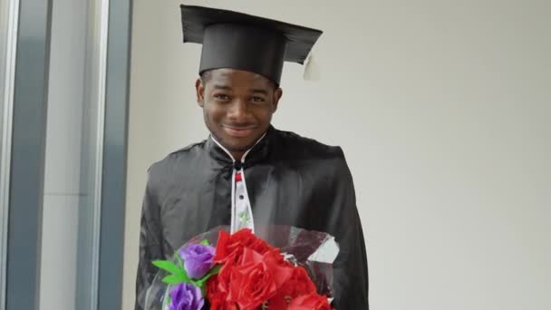 Um jovem afro-americano graduado em um manto e um chapéu mestrado e sorrindo, segurando um buquê de flores e posando para a câmera. Um evento importante. Jovem especialista — Vídeo de Stock