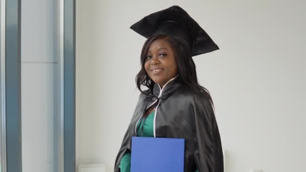 Una mujer afroamericana graduada con un traje y manto de maestría clásico tiene un diploma en sus manos y sonríe. Querido graduado de la Universidad Médica. Educación superior para mujeres en el extranjero — Vídeos de Stock