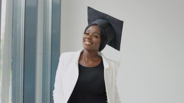 Una mujer afroamericana graduada con un lujoso traje blanco clásico y un sombrero máster lleva un diploma en las manos y sonríe. Educación superior para mujeres en el extranjero — Vídeo de stock