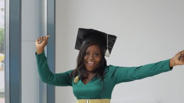 An African American female graduate in a green holiday dress with gold accessories and a square masters hat stands with a diploma in her hands and dances. Higher education for women abroad — Stock Video