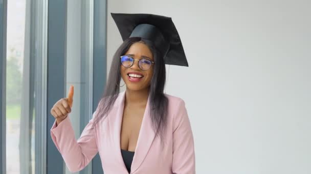 Una mujer afroamericana graduada con un traje clásico rosa claro y un sombrero maestro cuadrado posa y baila alegre y enérgicamente. .. Educación superior para mujeres en el extranjero — Vídeos de Stock