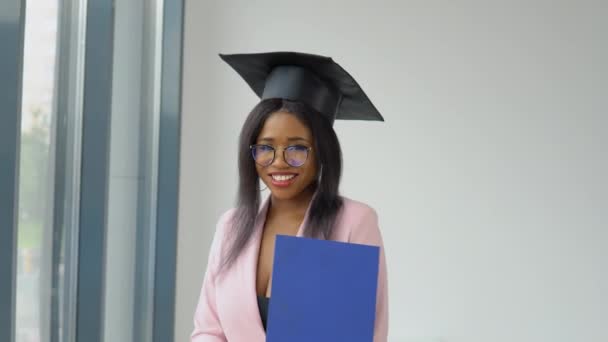 Афро-американская выпускница в светло-розовом классическом костюме и шляпе квадратного мастера с дипломом в руке весело и энергично танцует и позирует. Высшее образование для женщин за рубежом — стоковое видео