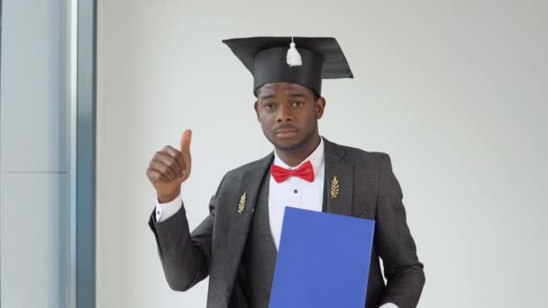 Um jovem afro-americano graduado em um chapéu de mestrado e um terno festivo cinza sorri, segurando um diploma e posando para a câmera. O início de um caminho profissional — Vídeo de Stock