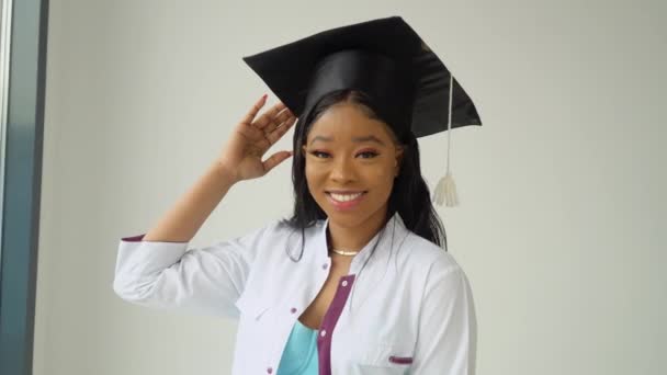 Une jeune femme afro-américaine diplômée en chapeau de maître et robe médicale blanche sourit pose pour la caméra. Un événement important. Jeune spécialiste — Video