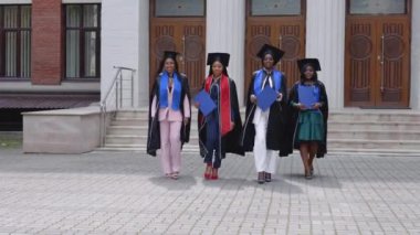 Afrika kökenli üniversite ya da üniversite mezunları ellerinde mavi diplomalarla üniversitenin girişinden kameraya doğru yürüyorlar.