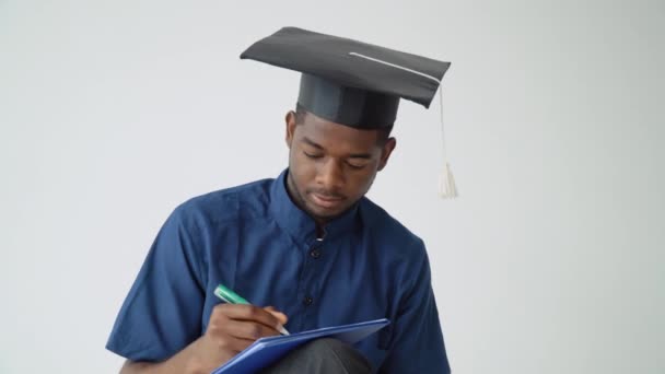 Un jeune homme afro-américain diplômé en chapeau de maître et blouse médicale bleue prend des notes. Un événement important. Jeune spécialiste — Video