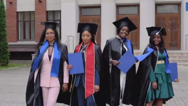 Graduados felizes de uma universidade ou faculdade de nacionalidade afro-americana com diplomas azuis em suas mãos posando na frente da universidade — Vídeo de Stock